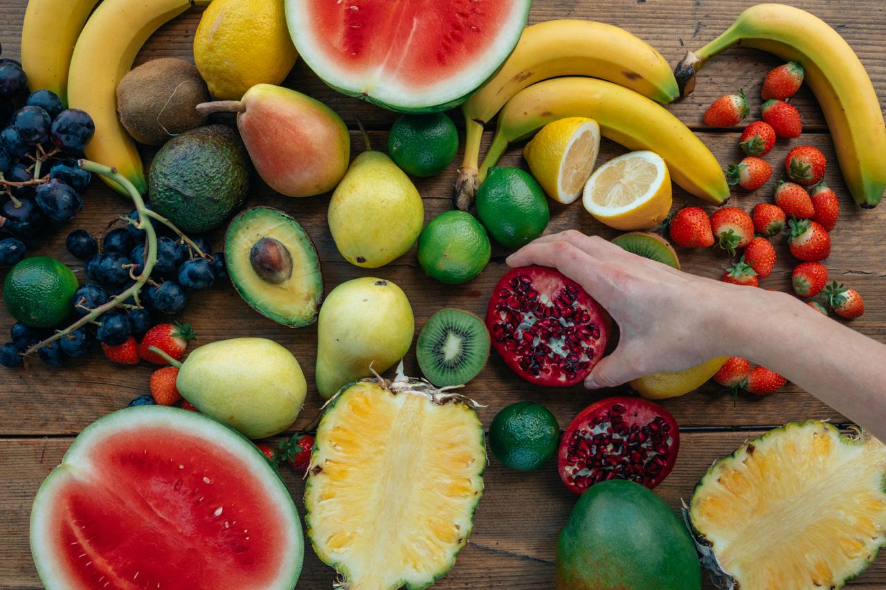 Colorful assortment of fresh fruits on wooden surface with a hand holding a pomegranate half.
